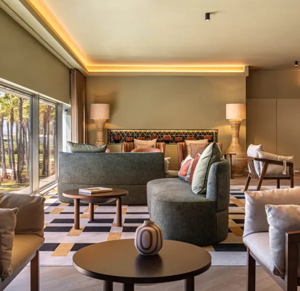 A modern, stylish hotel suite living area with round green and beige seating, a geometric rug, and large windows overlooking palm trees.